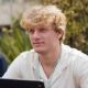 Young man with curly blond hair, wearing a white shirt, outdoors with greenery in the background.
