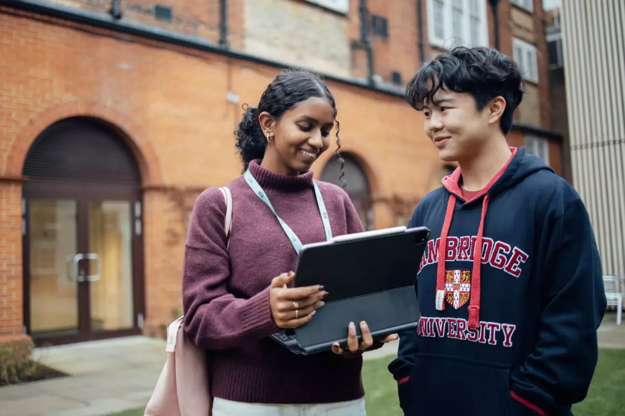 A pair of students, one male and one female, are standing and looking at a tablet while they chat.