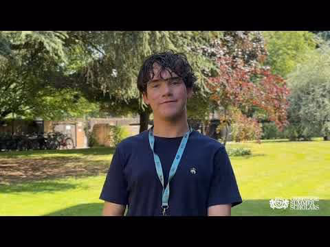Young man with dark curly hair wearing a dark shirt and light blue lanyard standing outdoors on a sunny day with trees and grass in the background.
