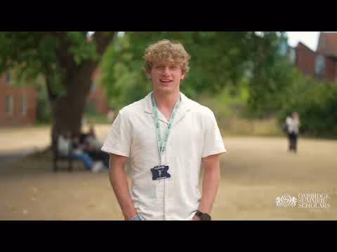 Young man with curly blond hair standing outdoors in a green park setting, wearing a white shirt and a lanyard with a badge.
