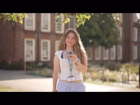 Smiling young woman wearing a white tank top and lanyard, holding a microphone outdoors with a brick building and trees in the background.