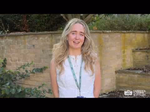 Smiling young woman with long blonde hair wearing a white sleeveless blouse and a lanyard, standing outdoors in front of a stone wall.