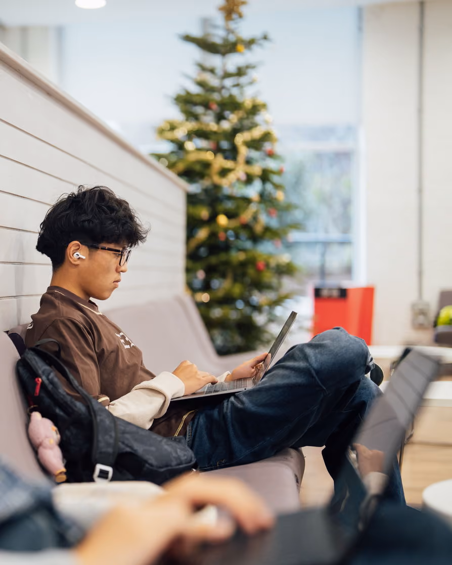 Person wearing glasses and earbuds working on a laptop on a couch with a decorated Christmas tree in the background.
