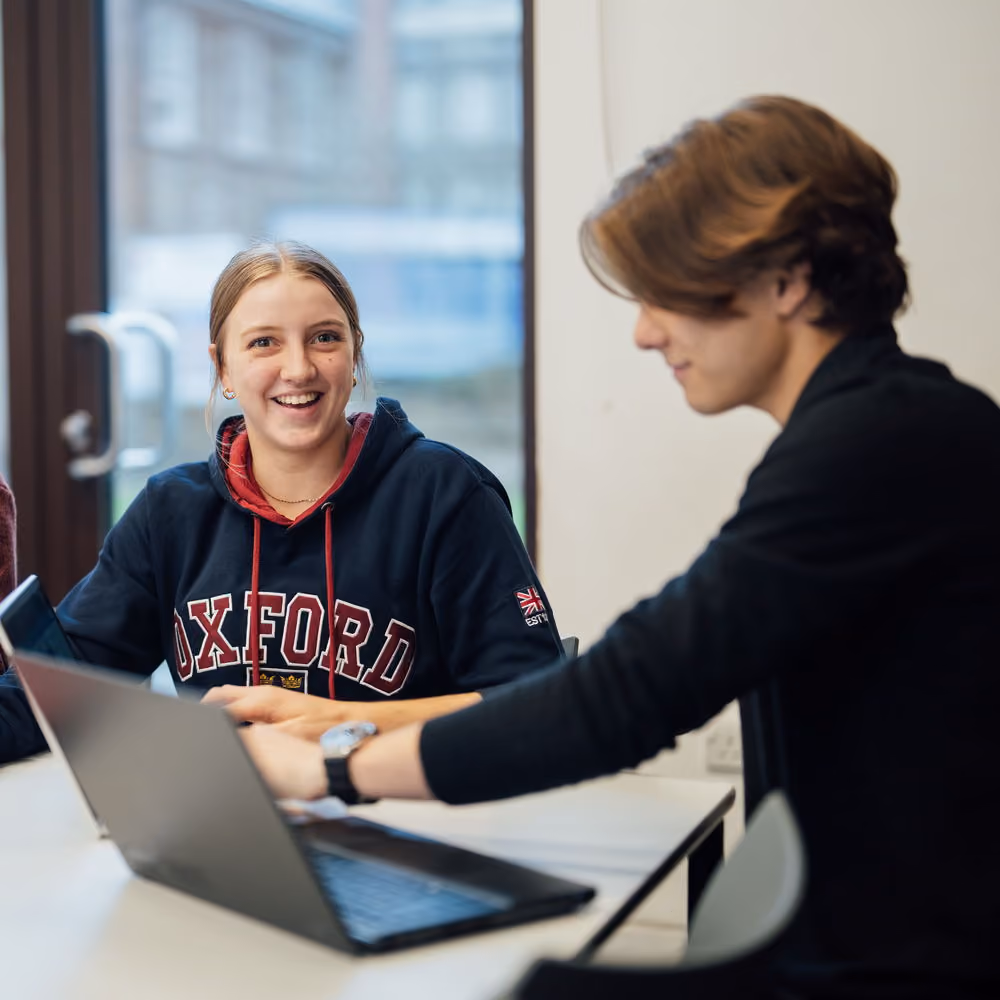 Two students working together at a table, one smiling and the other using a laptop.