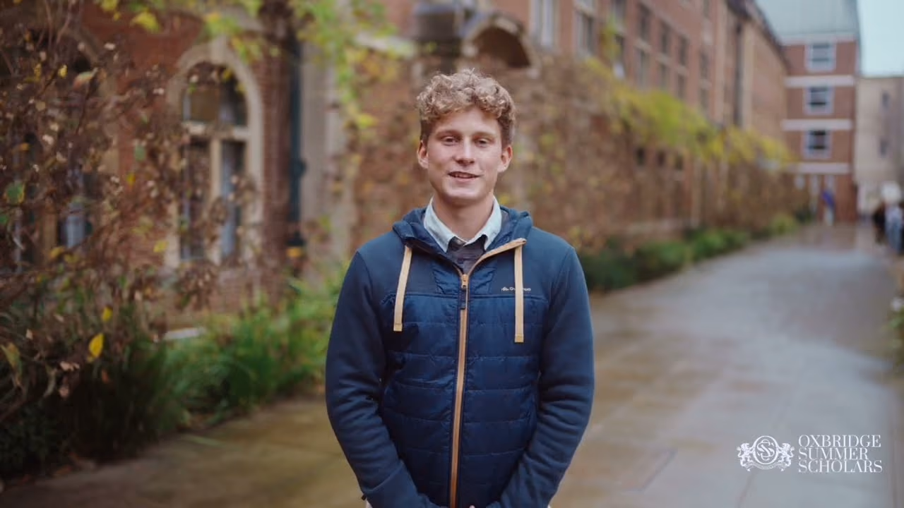 Young man wearing a black sweater standing outdoors near a brick building, holding a microphone and smiling.