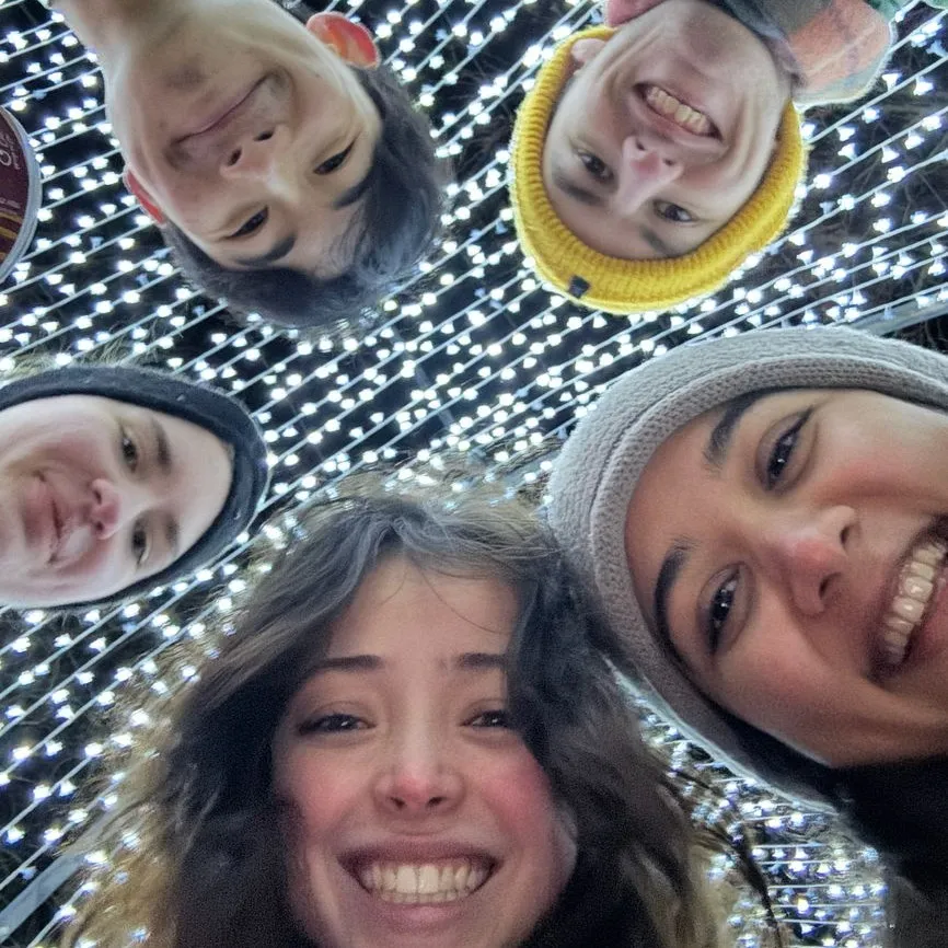 Five friends smiling in a close circle selfie with bright white string lights above them.