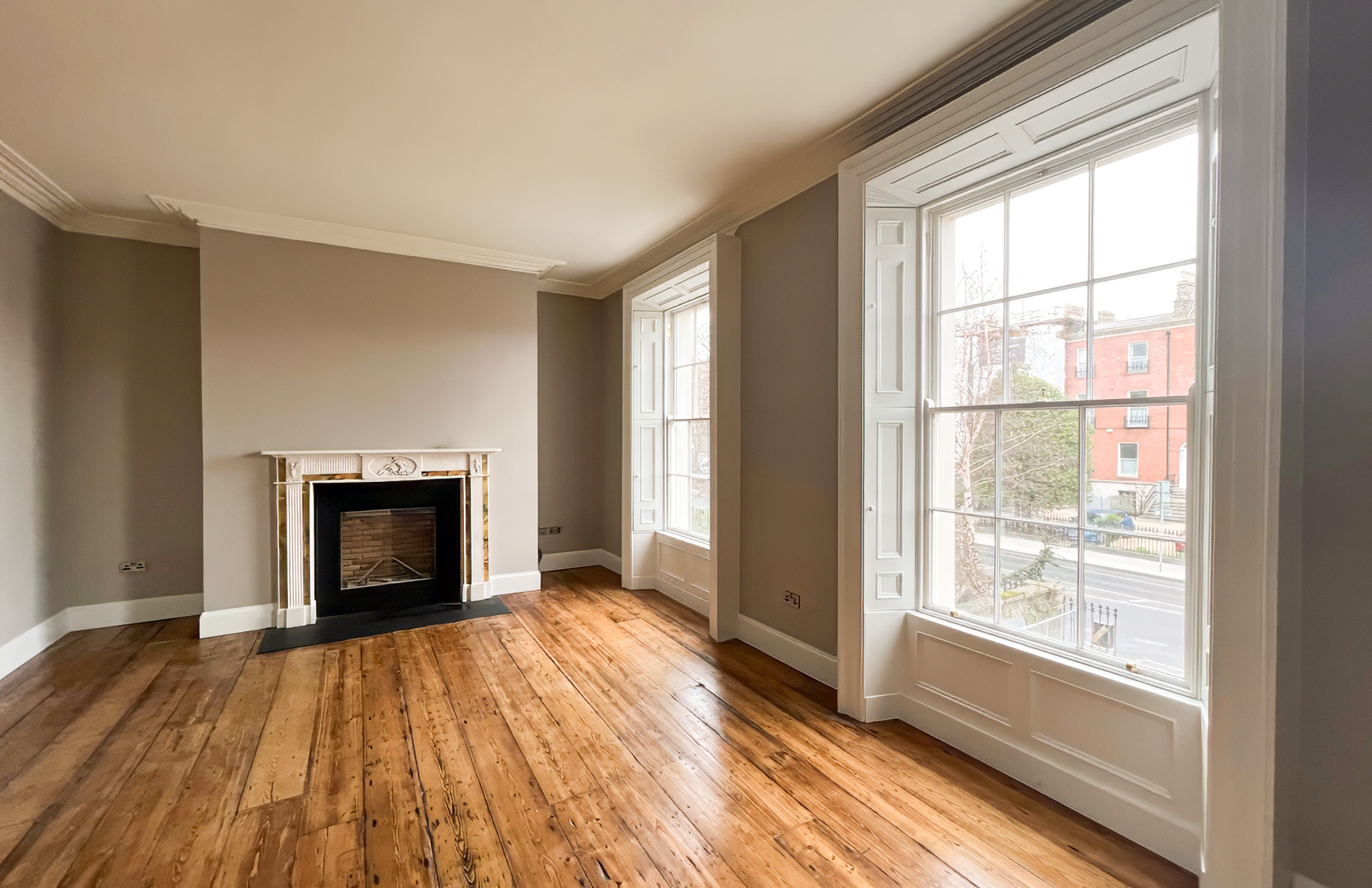 Modern living room with large windows, beige sofa, wooden chair, coffee table, indoor plants, and dining area in the background.