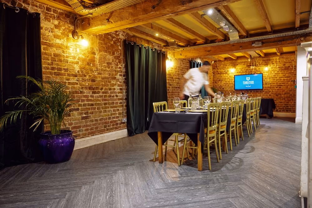 Long dining table set with glasses and plates in a brick-walled room with wooden beams and potted plant.