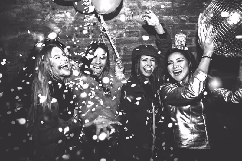 Four women laughing and celebrating with confetti, balloons, and a disco ball at a party.