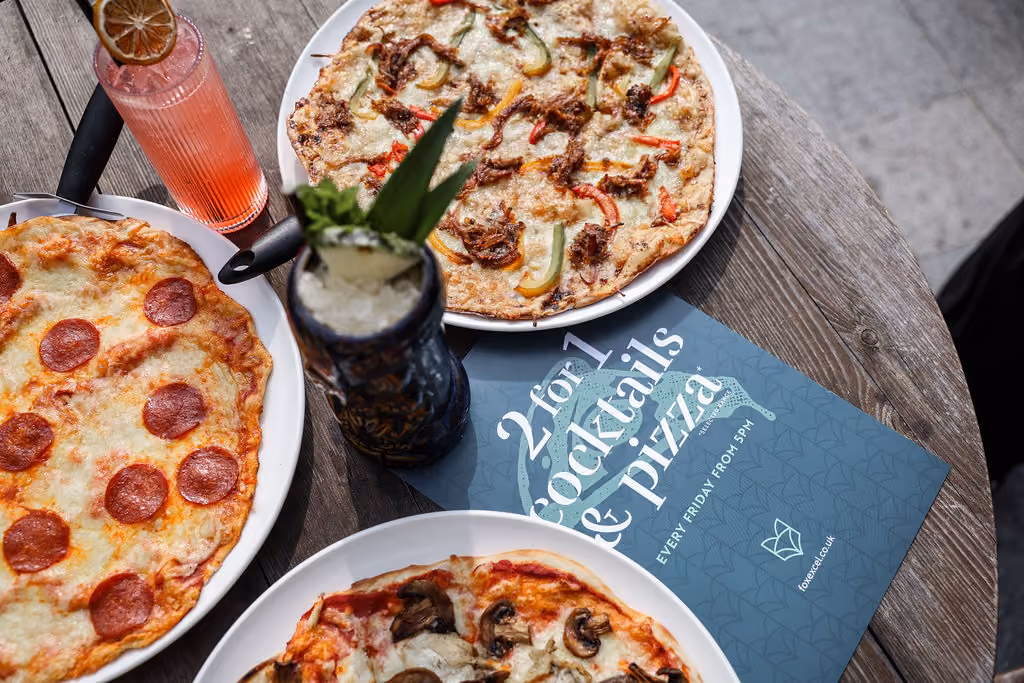 Table with three pizzas, two cocktails, and a blue sign advertising 2-for-1 cocktails and pizza Friday deal.