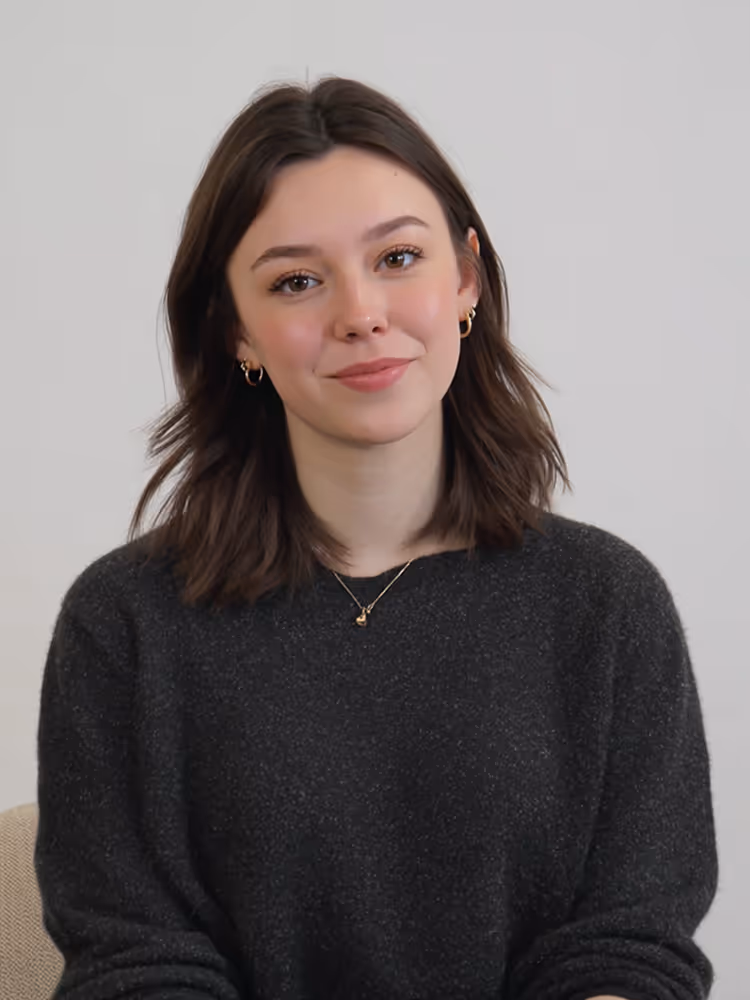 Young woman with shoulder-length brown hair wearing a dark gray sweater and small gold hoop earrings, smiling gently.