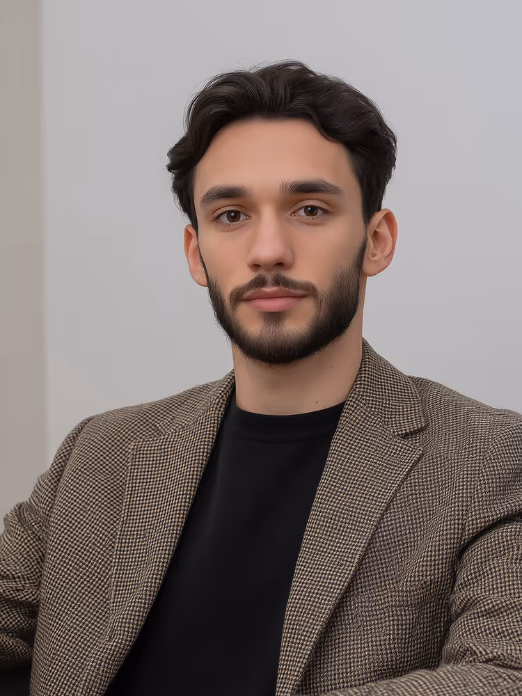 Portrait of a young man with dark hair and beard wearing a brown checkered blazer and black shirt against a plain background.