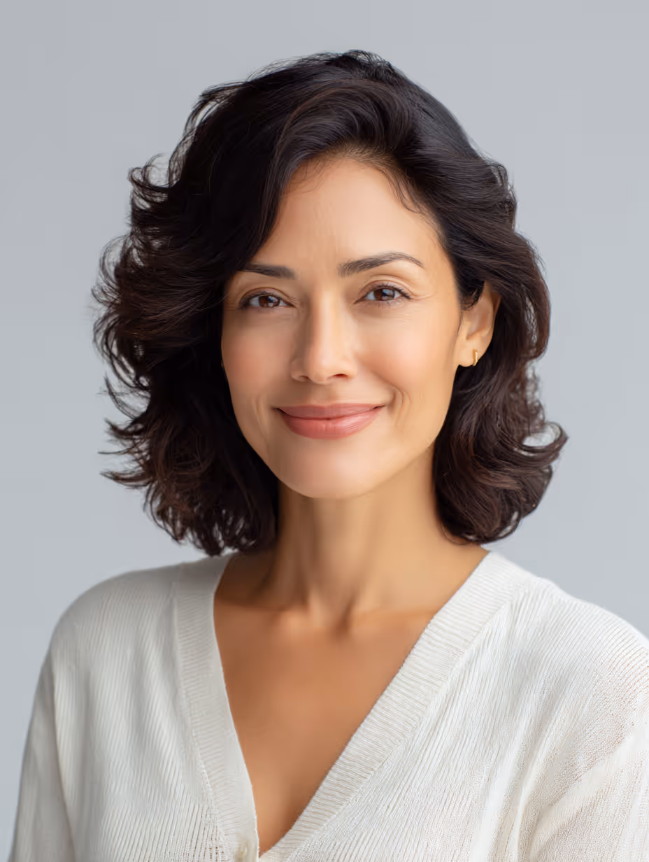 Smiling woman with shoulder-length dark wavy hair wearing a white cardigan against a light gray background.