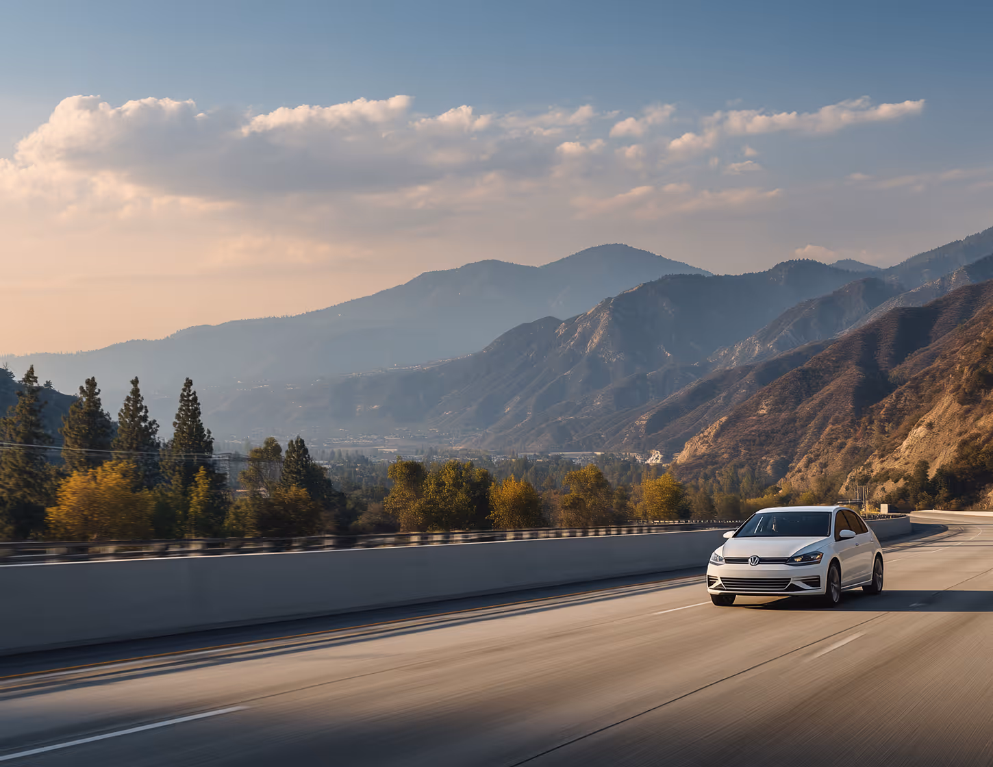 White car driving on a highway with mountains and trees in the background under a partly cloudy sky.