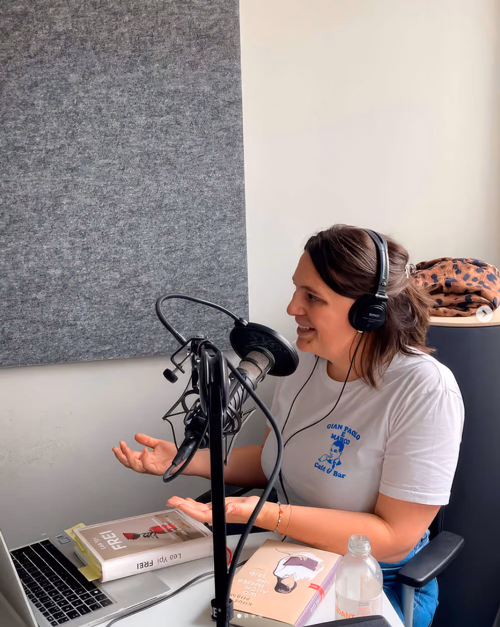 Annik Hosmann mit Kopfhörern spricht in Mikrofon, sitzt an Tisch mit Laptop, Büchern und Wasserflasche.