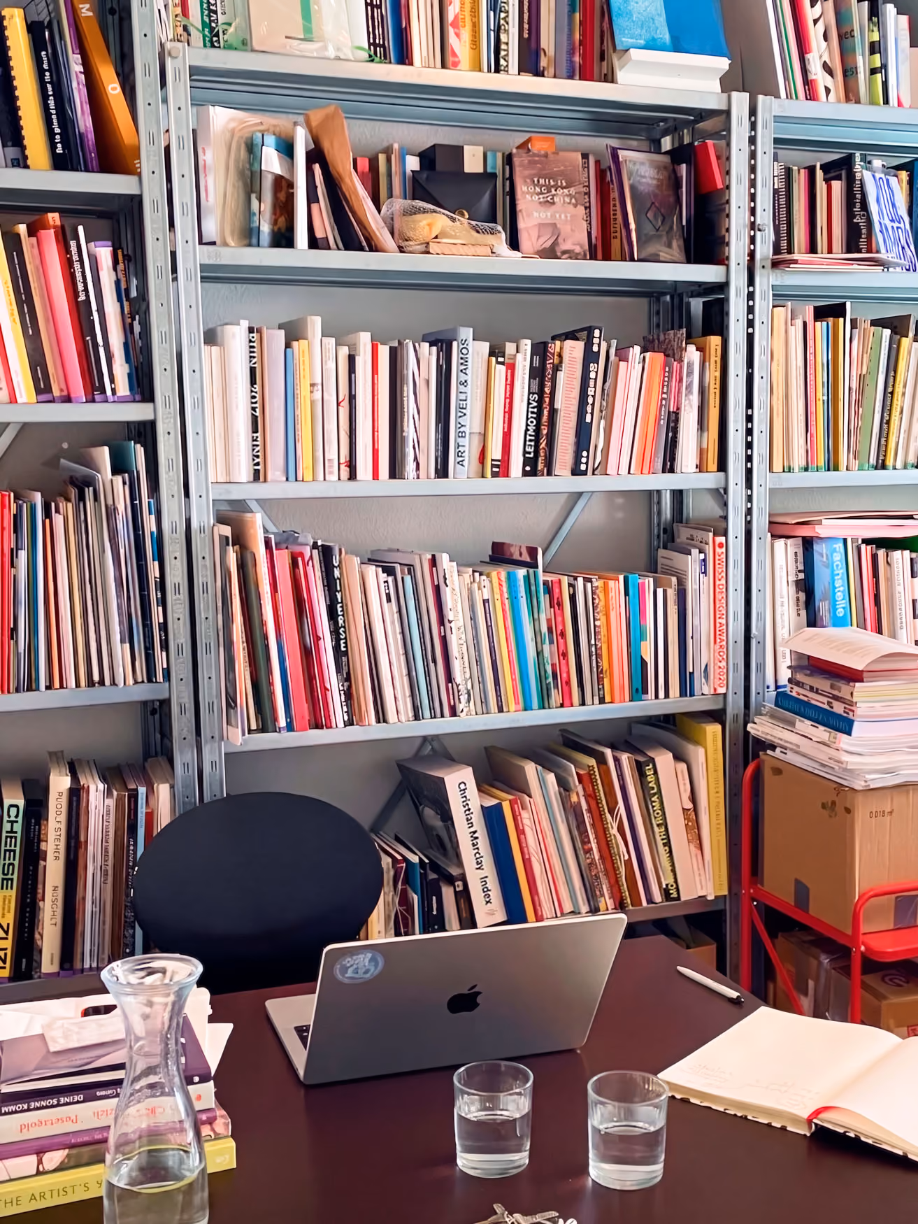 Wohnliches Büro mit vollgefülltem Bücherregal, Arbeitsplatz mit MacBook, Gläsern und offenem Notizbuch.