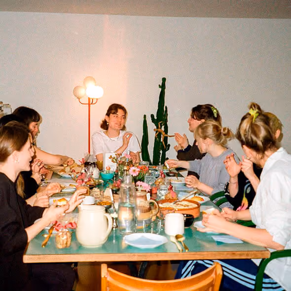 Menschen sitzen an einem Tisch und essen gemeinsam in einem hellen Raum mit einer Kaktuspflanze.