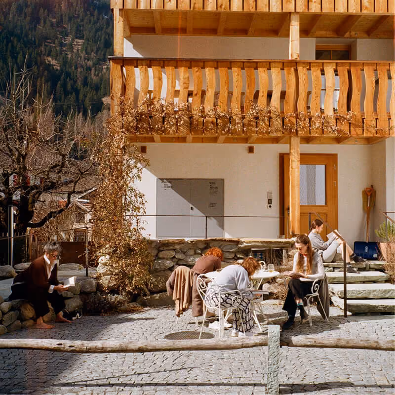 Menschen lesen im Sonnenlicht vor einem Holzhaus mit Balkon in einer bergigen Landschaft.