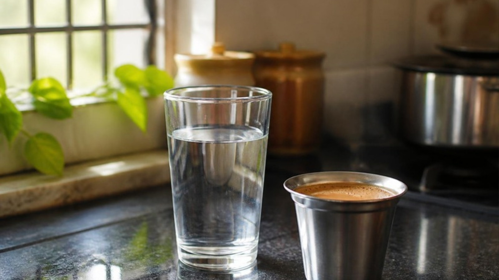 A glass of water on the kitchen counter beside a steel cup of chai