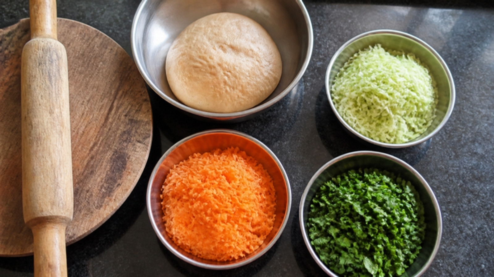 Grated carrot, palak and lauki in steel bowls beside paratha dough