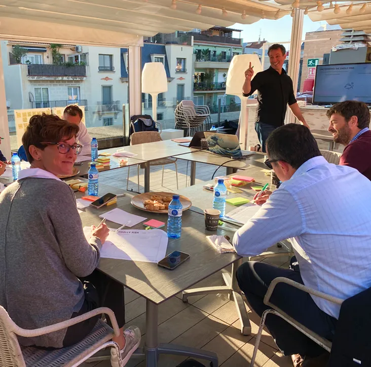 Group of people seated around a table on a sunlit terrace, engaging in discussion and taking notes with documents and water bottles on the table.