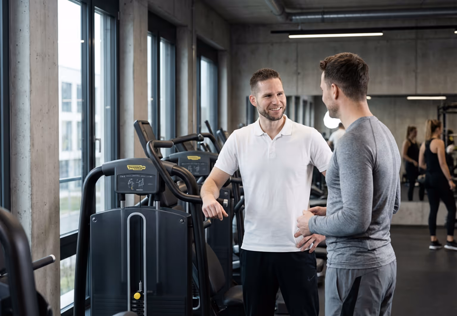Two men talking and smiling inside a modern gym with exercise machines and large windows.
