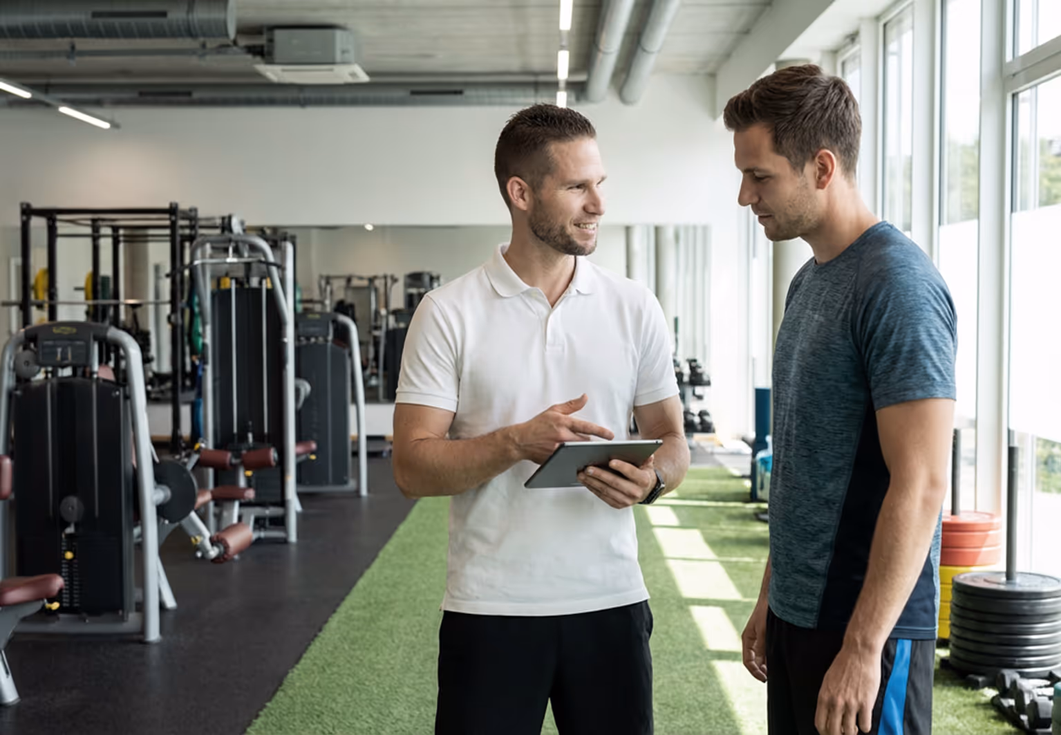 A fitness coach in a white polo shirt shows a tablet to a man in a blue workout shirt in a gym with exercise machines and weights.