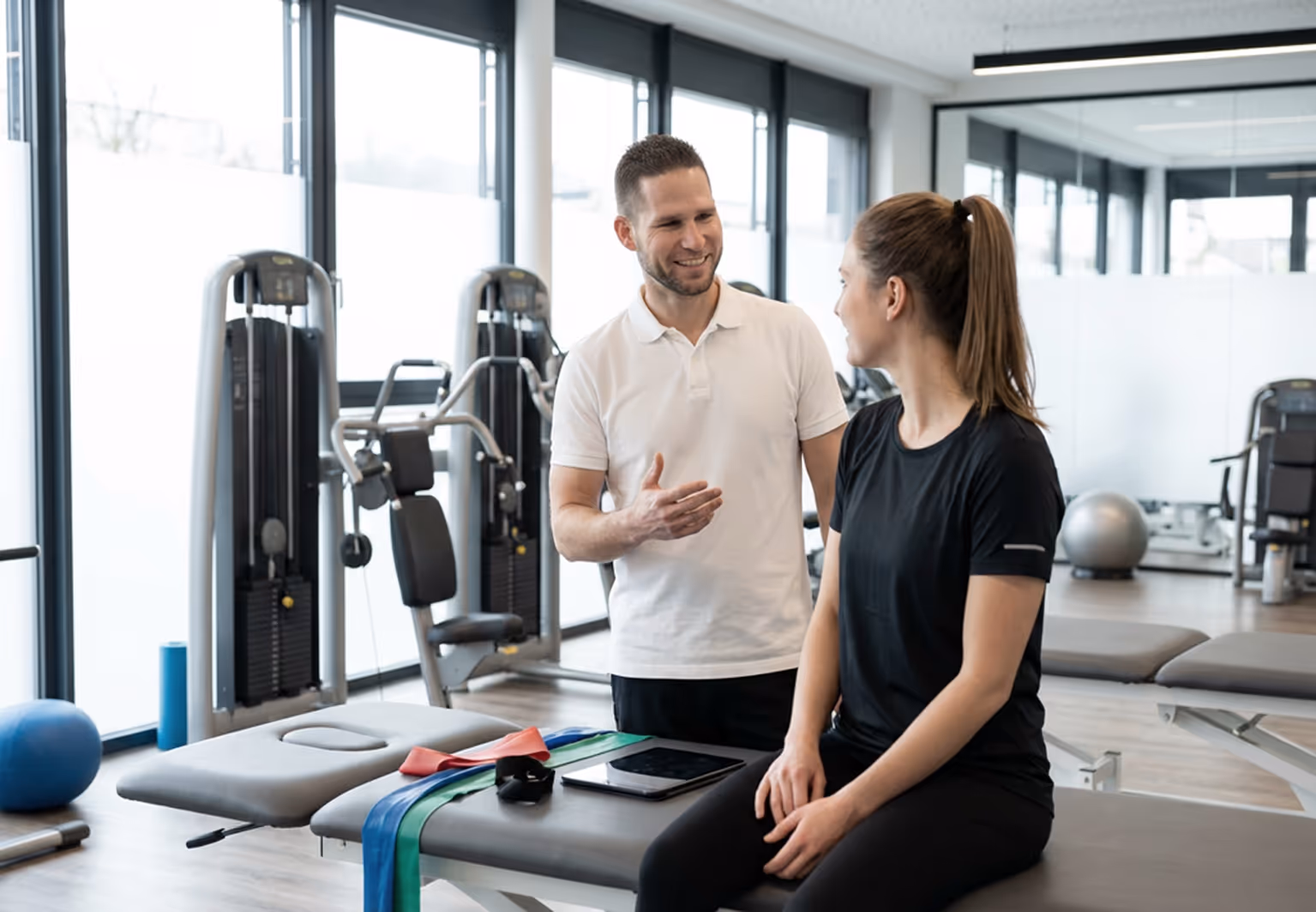 A male fitness trainer in a white shirt talks to a woman in black workout clothes sitting on a gym bench with resistance bands and a tablet beside her.