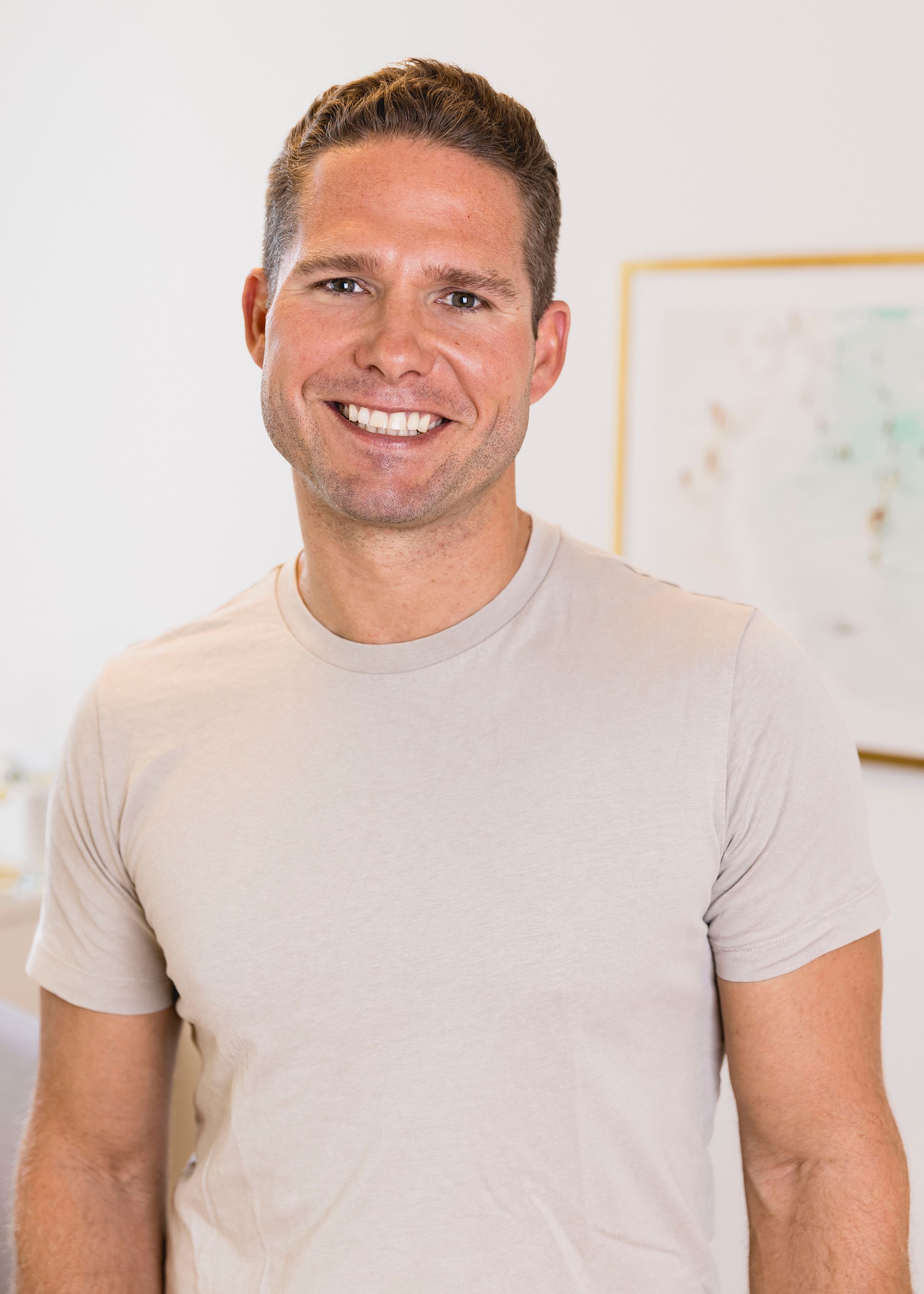 Smiling man with short hair and beard wearing a white polo shirt against a light peach background.