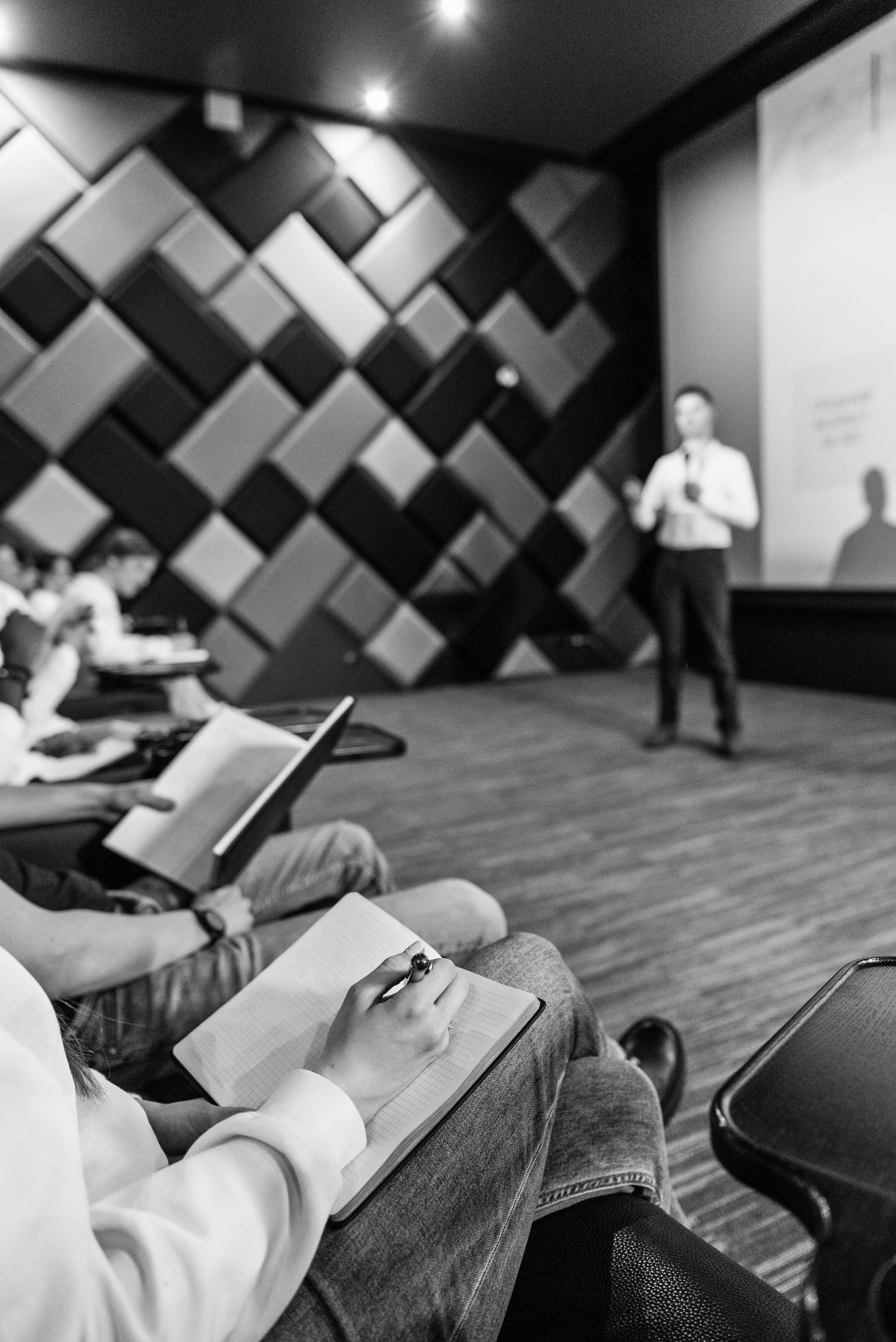Person taking notes in a notebook during a lecture or presentation in a modern conference room.