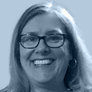 Smiling middle-aged woman with glasses, hoop earrings, and shoulder-length hair against a plain background.