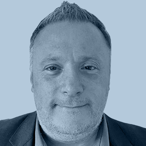 Portrait of a middle-aged man with short hair, light stubble, wearing a dark blazer and shirt, against a light blue background.