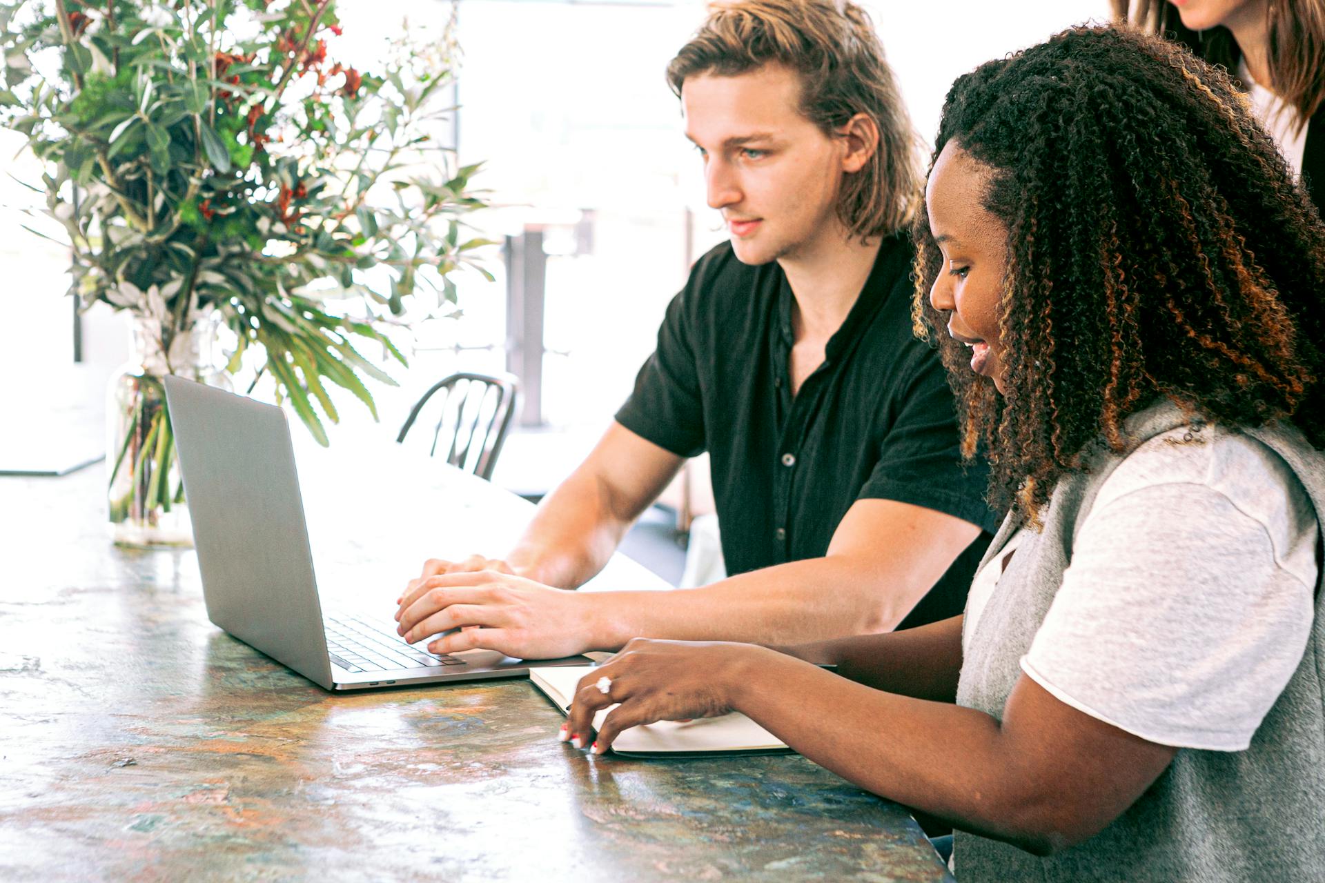 Two people working together on a laptop at a table with a large plant in the background.