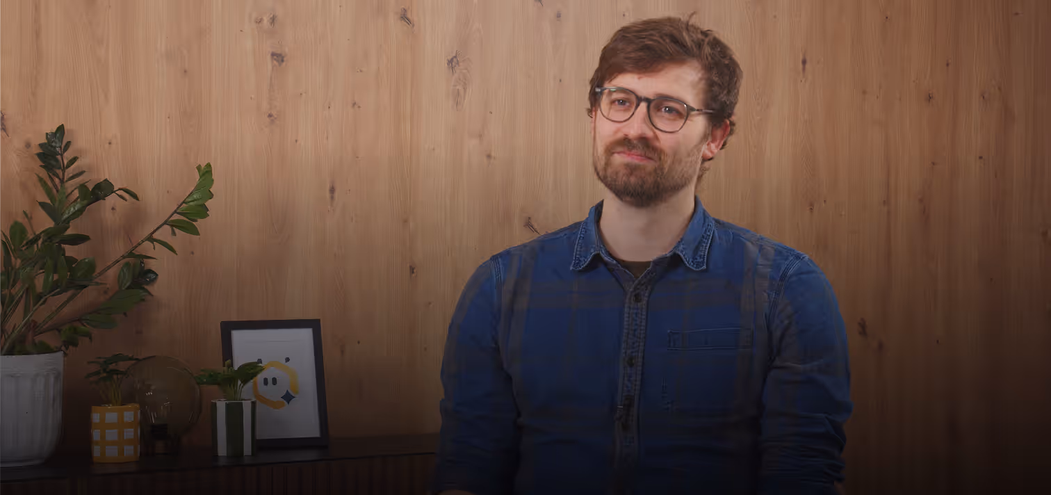 Man with glasses and beard wearing a blue shirt sitting in front of a wood-paneled wall with small plants and a framed picture on a sideboard.