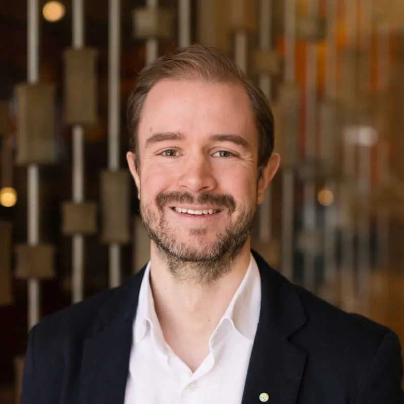 Smiling man with light brown hair and beard wearing a white shirt and dark blazer indoors.