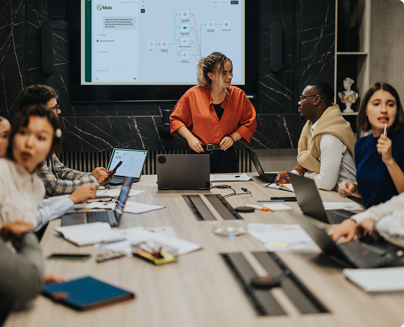 Diverse group in office meeting room with laptops, woman standing and speaking near a presentation screen.