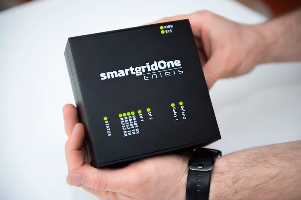 Hands holding a black smartgridOne Eniris device with labeled indicator lights for status, DI1, DI2, and relays.