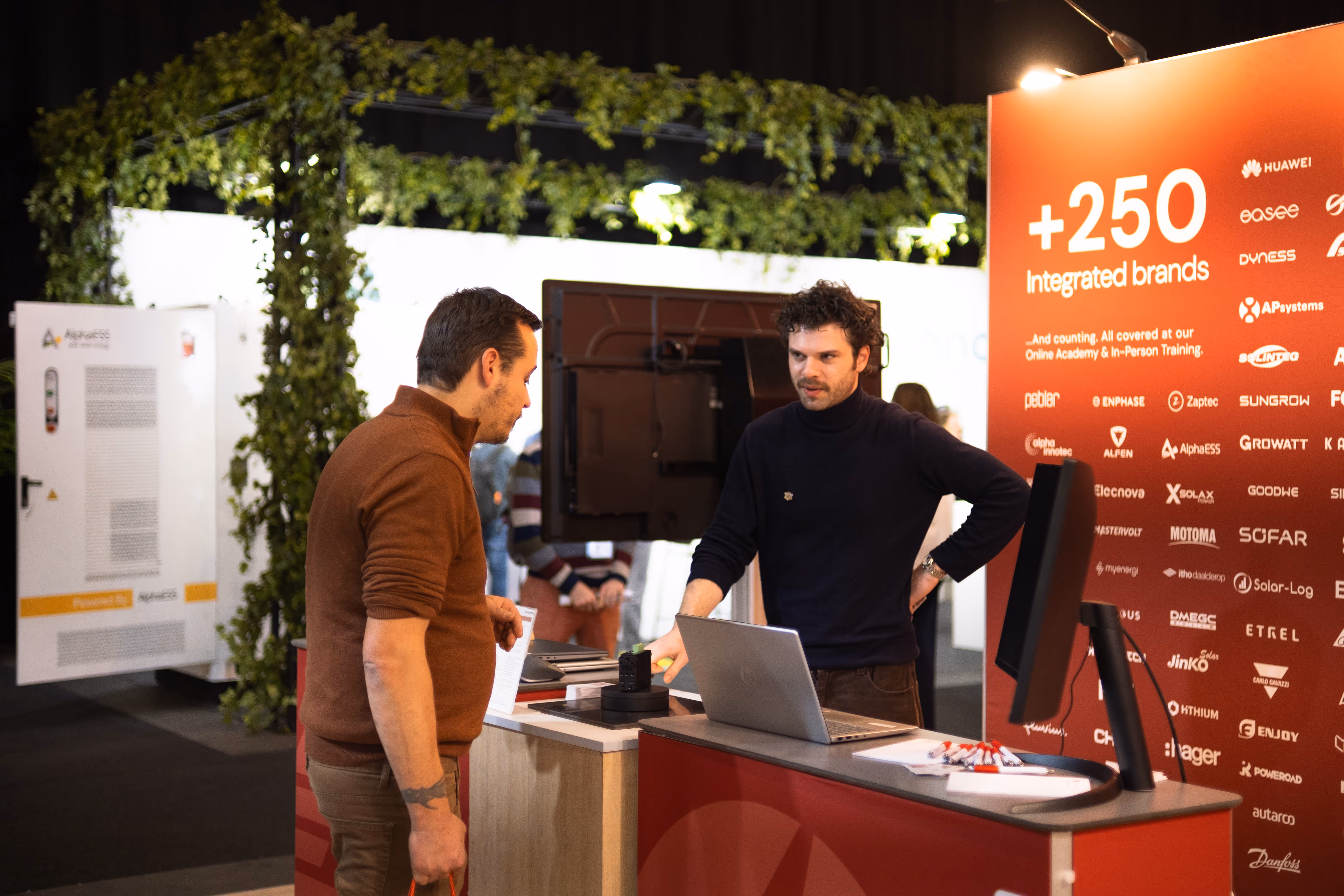 Two men conversing at a trade show booth with a laptop and a red backdrop listing over 250 integrated brands.