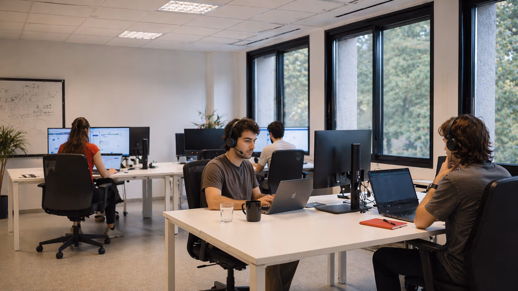 Four people working at desks with computers in an office with large windows and a whiteboard.
