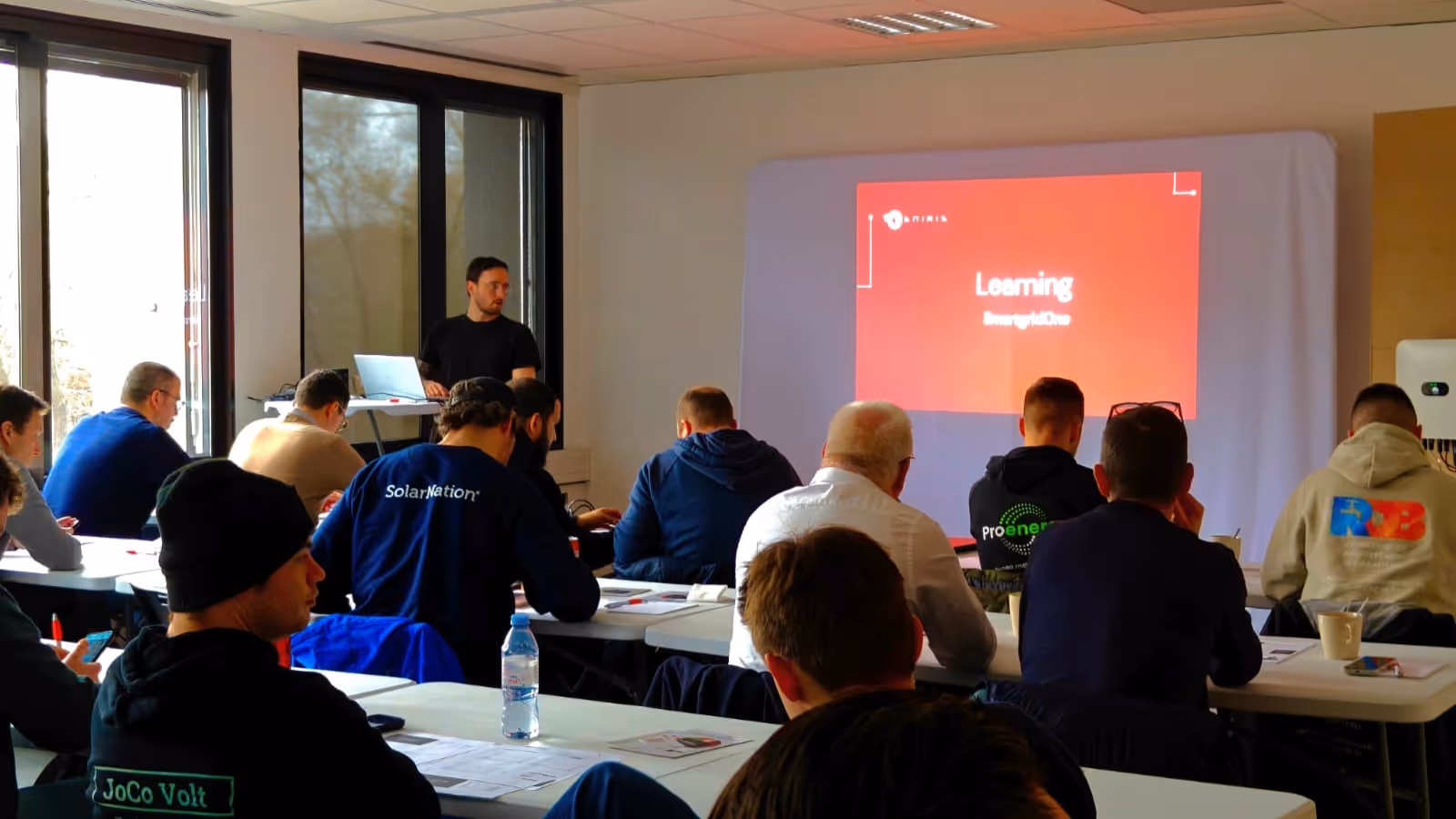 Instructor giving a training session to attendees seated at tables, with a red slide reading Learning projected on the wall.