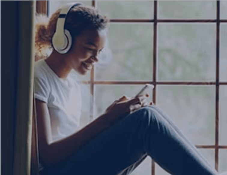 Jeune femme assise près d'une fenêtre, portant un casque audio et utilisant un smartphone.