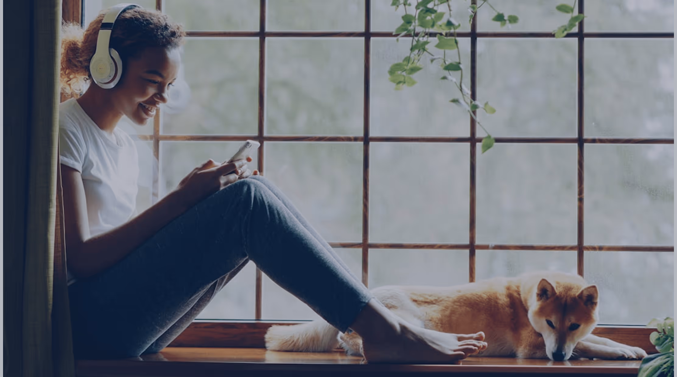 Jeune femme souriante avec des écouteurs, assise sur un rebord de fenêtre en bois, utilisant son téléphone, avec un chien allongé à côté d'elle.