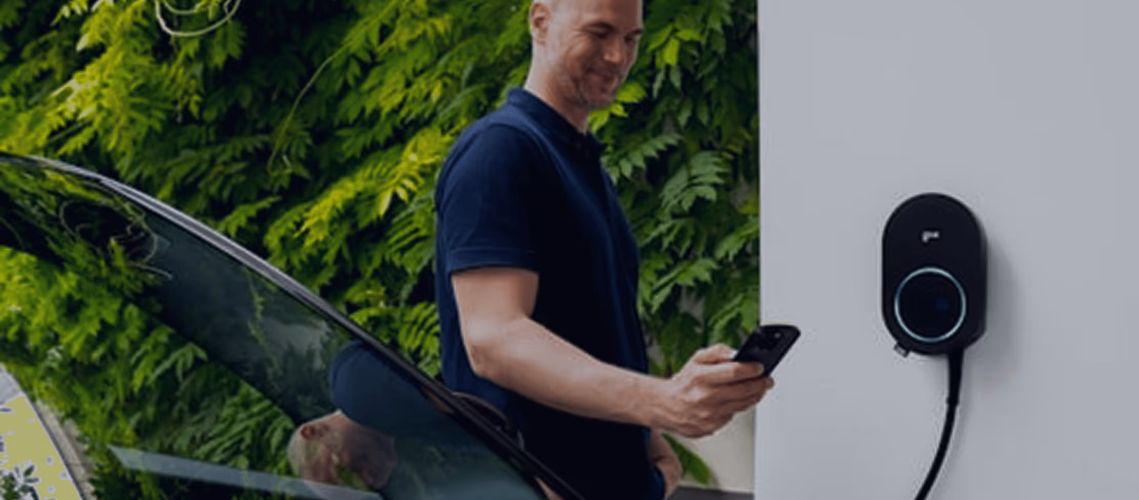 Un homme regarde son téléphone près d'une borne de recharge de voiture électrique installée sur un mur blanc, avec une végétation verte en arrière-plan.