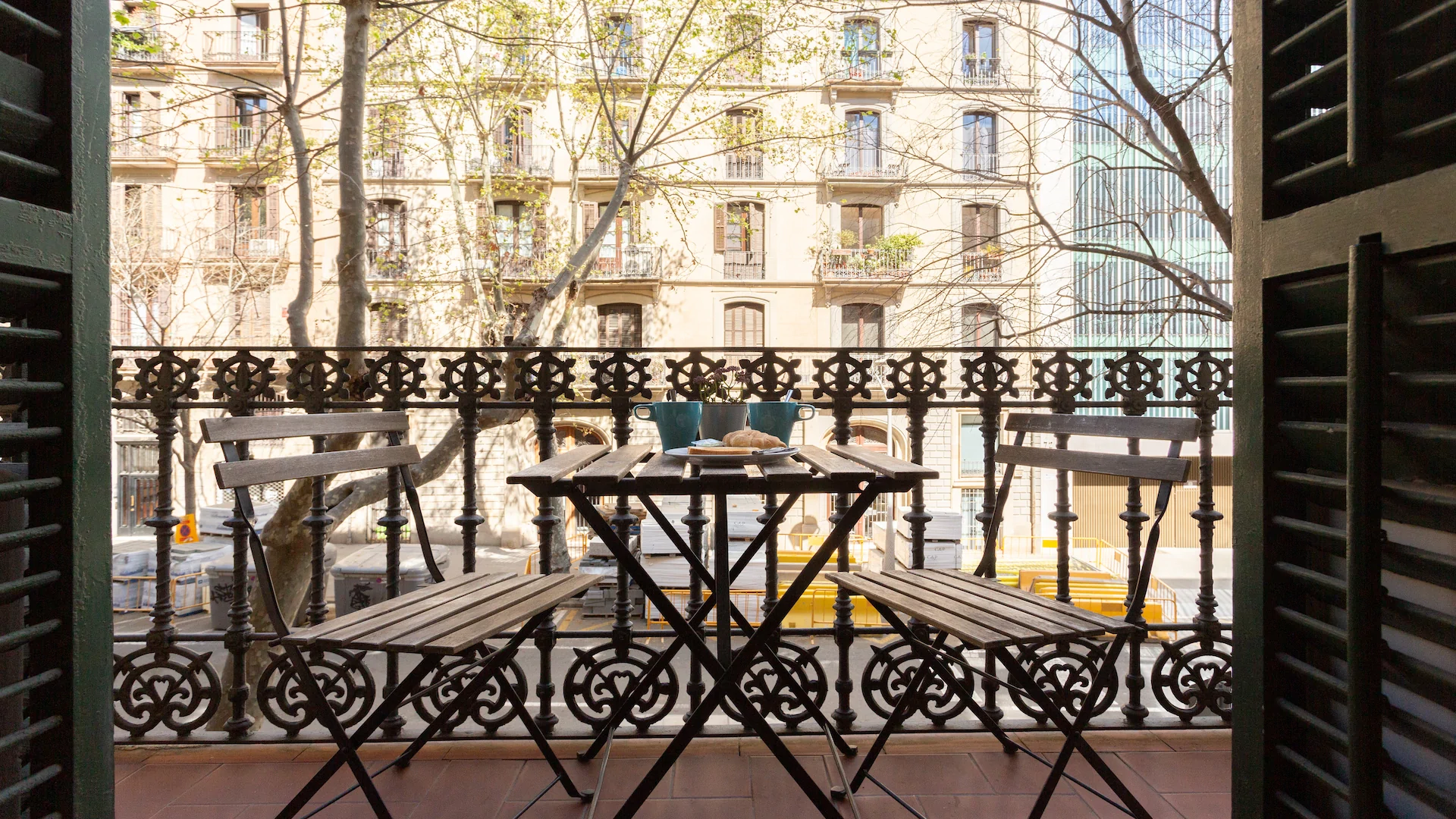 Small balcony with wooden table and two chairs, two blue mugs, and a plate with croissants, overlooking a sunlit street with buildings and trees.