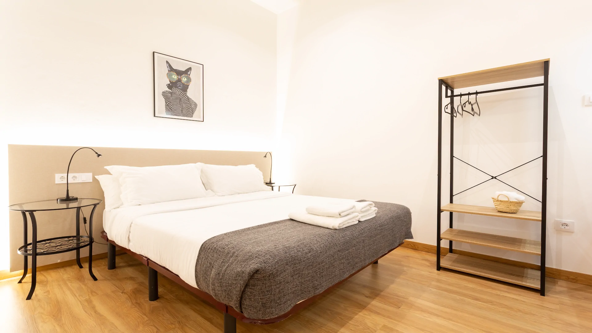 Minimalist bedroom with a double bed featuring white linens, a gray throw, bedside tables with lamps, a framed cat art on the wall, and an open wardrobe with hangers and a basket of towels.
