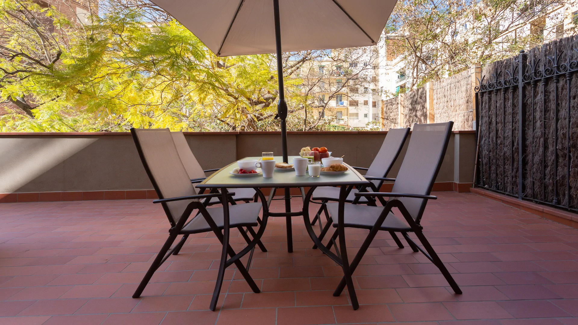 Outdoor patio dining table with four chairs under a large umbrella, set with breakfast items including fruit, croissants, and drinks, surrounded by trees and buildings.