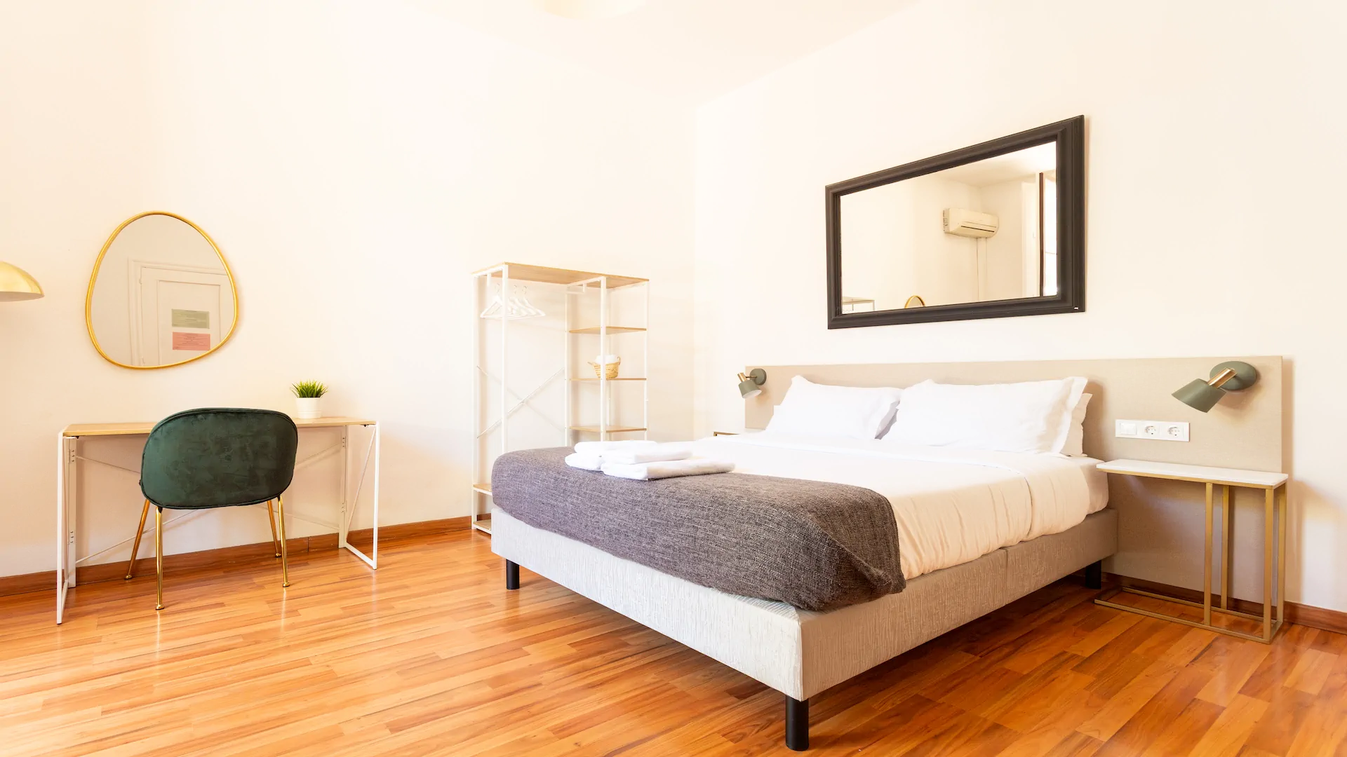 Minimalist bedroom with wooden floor, a bed with white linens and gray blanket, green chair at desk, and large rectangular mirror above the bed.