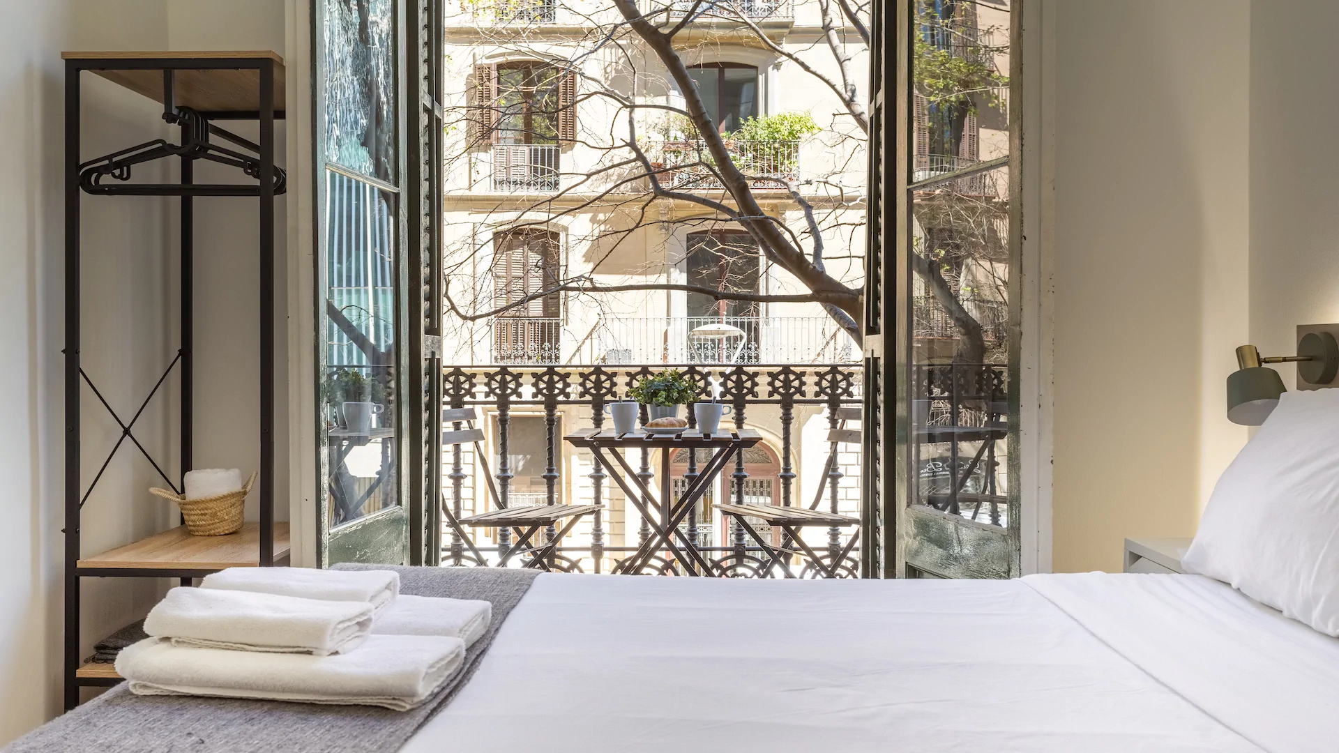 Minimalist bedroom with a neatly made white bed facing open glass doors leading to a balcony with a small table, chairs, and potted plant.