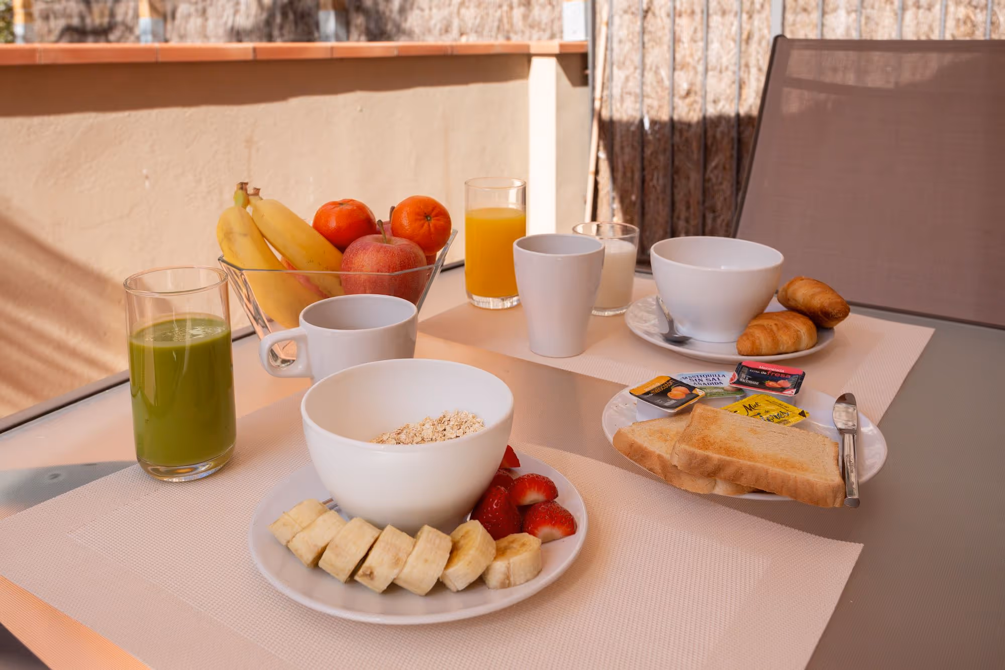 Breakfast table with sliced banana, strawberries, oats, toast with spreads, croissants, banana, apple, oranges, green juice, orange juice, and cups of coffee and milk.