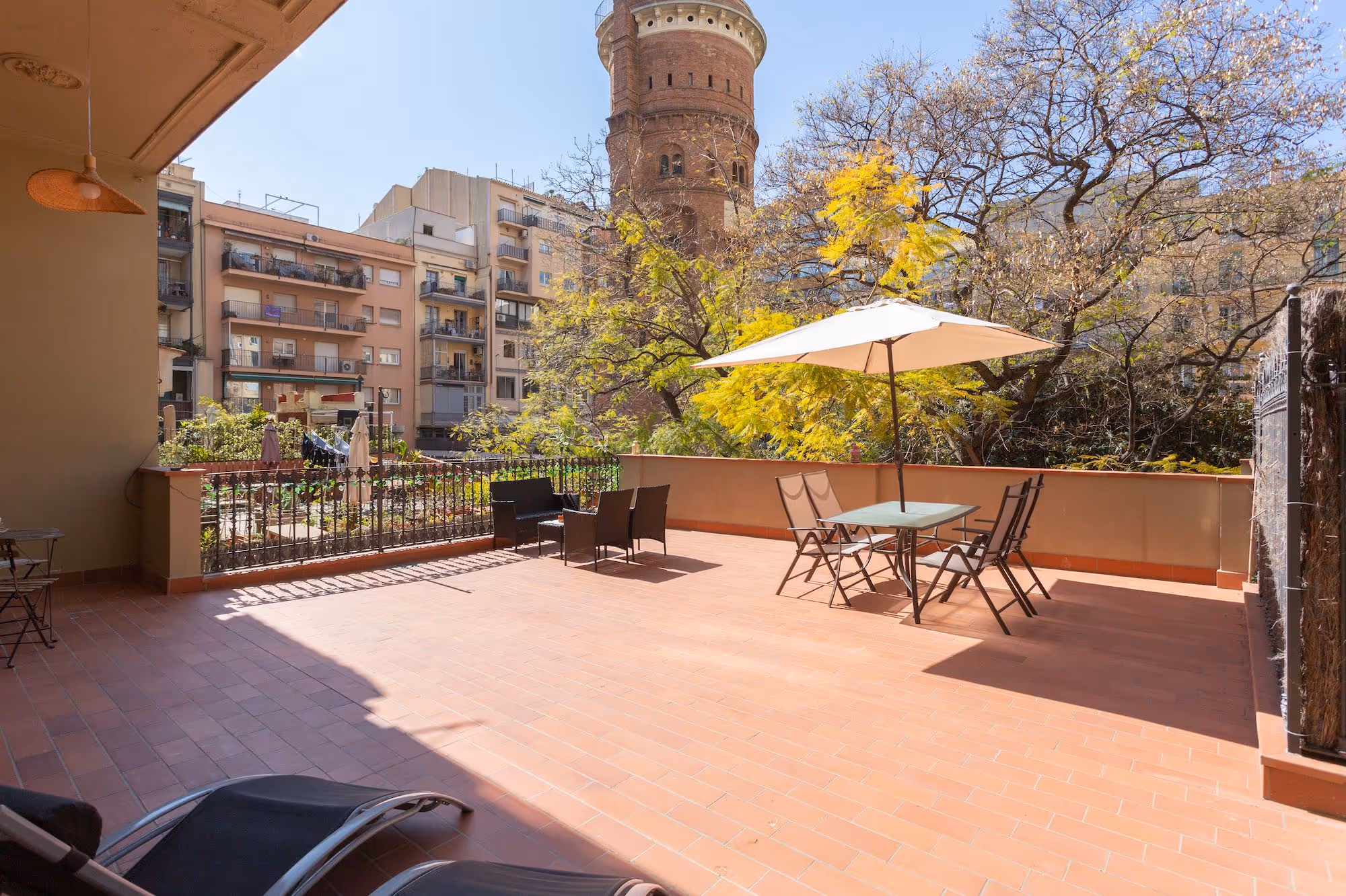 Spacious outdoor terrace with tiled floor, metal chairs, table with umbrella, and a view of an old brick tower and nearby apartment buildings.